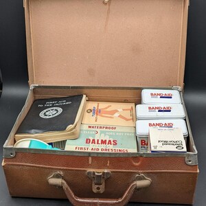 May include: A vintage brown leather first aid kit, open to show medical supplies. Contains Band-Aids, Dalmas first-aid dressings, and a booklet titled "First Aid to the Injured."