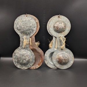 Puede incluir: Dos juegos de címbalos de mano antiguos de metal, cada juego consta de tres címbalos conectados por un cordel. Los címbalos son de color gris desgastado con manchas de óxido.