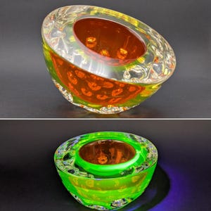 Puede incluir: Un cuenco de vidrio grueso y transparente con forma redondeada y asimétrica. El interior presenta una cuenca circular de color naranja intenso. Bajo luz ultravioleta, la capa exterior de vidrio brilla en un color verde neón vibrante.