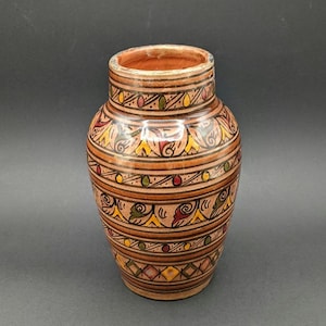 May include: A terracotta vase with a glossy finish, featuring horizontal bands of hand-painted geometric and floral patterns in yellow, red, and green. The vessel has a wide neck, a rounded body, and a flat base, showcasing intricate line work.