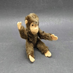 Könnte beinhalten: Ein kleiner, brauner Plüschaffe mit hellbraunem Gesicht, Händen und Füßen. Der Affe hat ein strukturiertes Fell und ist mit ausgestreckten Armen posiert. Das Spielzeug ist im Vintage-Stil und wahrscheinlich ein Sammlerstück.