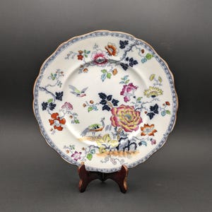 Puede incluir: Un plato decorativo vintage con borde festoneado, con un diseño floral y de aves. El plato blanco presenta flores coloridas en rosa, naranja y amarillo, y un pavo real y un pájaro. El borde tiene un borde azul y dorado.