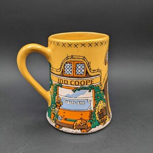 May include: A yellow ceramic mug featuring a colorful illustration of a pub exterior. The design includes the text Ind Coope and Public Bar, a thatched roof, green vines, and an orange cat resting on a wooden bench near the base of the mug.