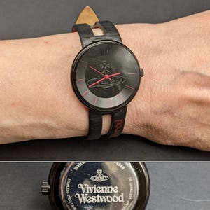 May include: A black wristwatch with a round face featuring a red orb logo and red hour markers. The watch has a black leather strap with a split design and a stainless steel back engraved with the Vivienne Westwood logo and text.