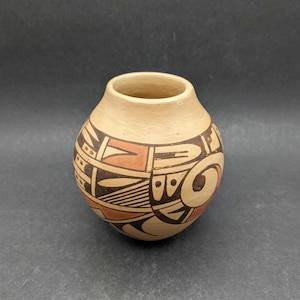 May include: A small, round, tan-coloured clay pot featuring a wide opening at the top. The exterior surface is decorated with intricate geometric patterns in black and terracotta-coloured paint, including swirling lines, triangles, and dots.