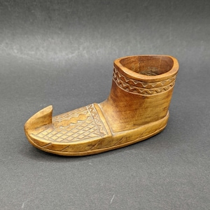 Puede incluir: Un adorno de madera con forma de zapato con intrincados tallados. El zapato está hecho de madera marrón claro y tiene una pequeña abertura en la parte superior.