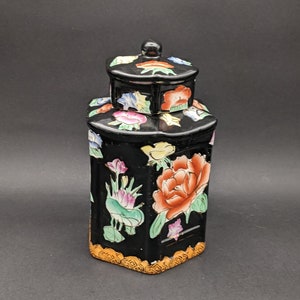 Peut inclure: Un pot en porcelaine noire avec un couvercle, décoré de motifs floraux colorés. Le pot a une forme hexagonale et une bordure dorée autour de la base.