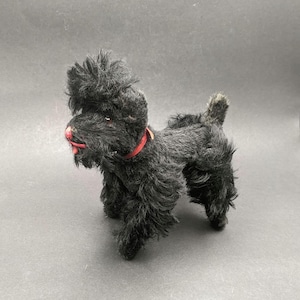 Puede incluir: Un perro de juguete negro vintage con un collar y una nariz rojos. El perro tiene un pelaje esponjoso y texturizado y está de pie sobre una superficie gris. El juguete es un caniche.