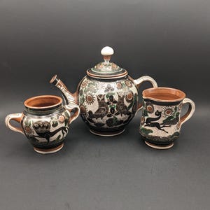 May include: A ceramic teapot and two mugs with a black, green, and white floral design. The teapot has a round body and a lid with a knob. The mugs have a rounded shape and a handle. The design features stylized birds and flowers.