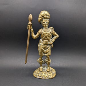 Puede incluir: Una figurita de latón de un soldado escocés con kilt y sosteniendo una lanza. La figurita está de pie sobre una base con una superficie texturizada.