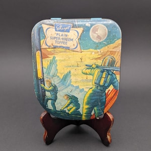 Może przedstawiać: Winietkowe pudełko z blachy z ilustracją kosmiczną przedstawiającą astronautów badających księżycowy krajobraz. Pudełko jest niebieskie i żółte z nadrukiem "Sharps Plain Super-Kreem Toffee" na przodzie.