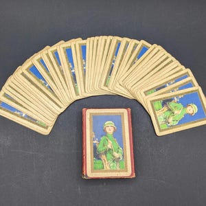 May include: Vintage deck of playing cards featuring illustrations of soldiers in green uniforms. The cards are fanned out, with a red and gold card case in the center. The cards have a blue and gold border.