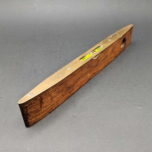May include: A spirit level tool featuring a dark brown wooden body with a polished brass top plate. The brass plate includes a central glass vial containing a bright yellow-green liquid and a bubble, secured by small metal screws at each end.