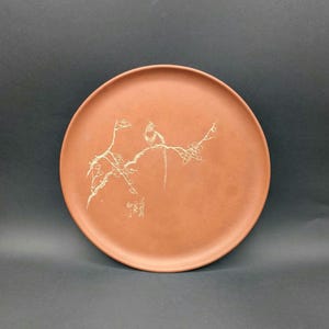 Puede incluir: Un plato redondo de color terracota con una ilustración detallada de un pájaro posado en una rama. El pájaro y la rama son de un color dorado más claro. El plato tiene una superficie lisa y un diseño simple y elegante.