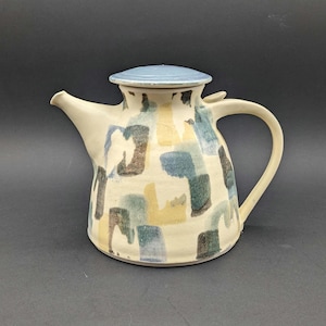 Puede incluir: Una tetera de cerámica con tapa azul claro y cuerpo color crema. El cuerpo está decorado con un patrón de formas azules, amarillas y marrones. La tetera tiene un asa curva y un pico.