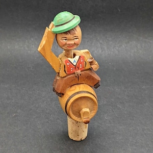 May include: Wooden bottle stopper with a carved figure of a man in a green hat and vest, sitting on a small barrel. The figure is holding a small cup. The stopper is made of wood and cork.