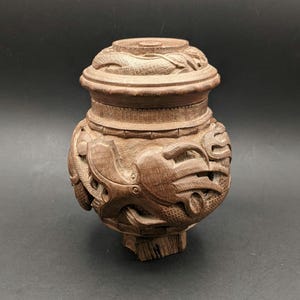 May include: A brown, carved wooden container with a lid. The container features detailed carvings of fish or sea creatures. The lid has a detailed pattern. The container sits on a small wooden base. The measurements are in centimetres.