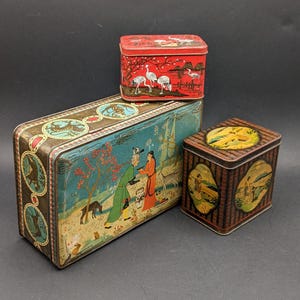 Può includere: Tre scatole di metallo vintage con disegni dipinti. La scatola più grande è rettangolare con una scena paesaggistica blu e marrone. Una scatola rossa più piccola presenta gru bianche. La scatola più piccola è marrone con disegni ovali gialli e neri.