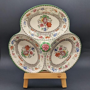 Könnte beinhalten: Eine dreigeteilte Keramik-Servierschale mit cremefarbenem Untergrund, verziert mit einem bunten Blumenmuster in Rot, Orange, Blau und Grün. Die Schale hat einen grünen Rand und einen mittigen Griff, der mit einer modellierten rosa Rose gekrönt ist.