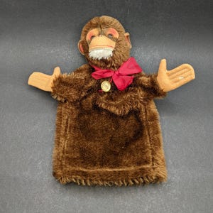 May include: A vintage brown monkey hand puppet with a red bow tie and tan hands. The puppet has a plush, furry texture and a detailed face with orange accents. A gold-colored medallion is attached to the bow.