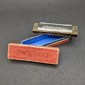 May include: A silver-toned metal harmonica with the word PICCOLO engraved on the top, resting above its open rectangular box. The box features a textured red exterior with the word PICCOLO printed on the lid and a bright blue interior lining.