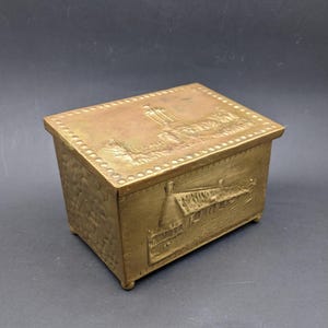 Puede incluir: Una caja de latón con un diseño en relieve de un edificio en la tapa y una casa en la parte delantera. La caja tiene un borde de cuentas alrededor de la tapa.