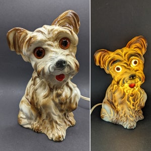 Puede incluir: Una lámpara de cerámica con forma de perro de pelo marrón y blanco, ojos marrones y lengua roja. La lámpara está encendida y tiene un brillo cálido.