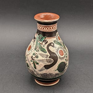 May include: A small, round, ceramic vase with a black, white, and green design. The vase features a stylized bird with a long neck and a floral pattern.