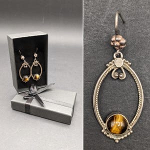以下が含まれることがあります： タイガーアイの石が中央にセットされた、シルバーカラーの楕円形イヤリング。 イヤリングは黒いリボンが付いた黒いギフトボックスに入っています。 片方のイヤリングが、濃いグレーの表面に別々に表示されています。