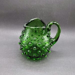 May include: A small, green glass jug with a textured, hobnail design. The jug has a rounded body, a handle, and a spout. The glass is a deep, emerald green colour. The hobnail pattern covers the entire body of the jug.