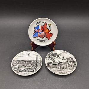 Könnte beinhalten: Drei kleine, weiße Souvenirteller mit Illustrationen von Berliner Wahrzeichen. Einer zeigt eine Karte mit Flaggen und dem Text "BERLIN CHECKPOINT CHARLIE". Die anderen beiden haben detaillierte Schwarz-Weiß-Zeichnungen von Gebäuden.