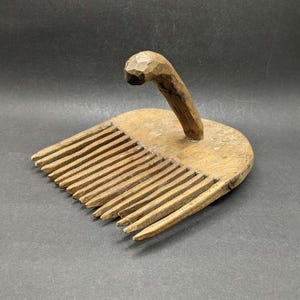 May include: A vintage wooden comb with a curved handle. The comb has a semi-circular base with multiple thin, evenly spaced teeth. The handle is attached to the base and has a carved, textured appearance. The wood has a natural, aged color.