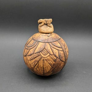 Può includere: Un contenitore in legno intagliato a mano con forma rotonda e intricati motivi floreali. Il coperchio è sormontato da una piccola statuina a forma di rana. Il contenitore è di colore marrone caldo e sembra essere fatto di legno naturale.