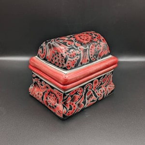May include: A black and red ceramic trinket box with a floral design. The box has a hinged lid and a red trim around the edges.