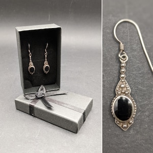 以下が含まれることがあります： 黒い楕円形の石が付いたシルバーカラーのドロップイヤリング。黒いリボンが付いた灰色のギフトボックスに入っています。イヤリングは、小さなビーズのアクセントが付いた装飾的なデザインです。