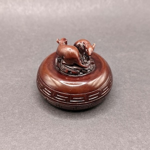 Könnte beinhalten: Eine kleine, runde Holzkiste mit einem Deckel, der zwei geschnitzte Ratten zeigt. Die Kiste ist dunkelbraun und hat ein dekoratives Muster am Rand.