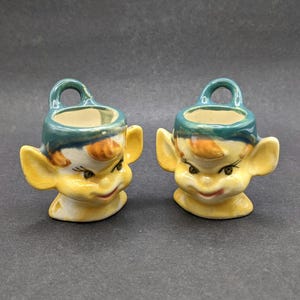 May include: A pair of ceramic egg cups shaped like elf heads with yellow skin, orange hair, and teal hats. Each cup features large ears, painted facial features including eyes and a mouth, and a small loop handle at the top of the teal hat.