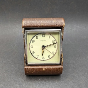 May include: A square Junghans travel alarm clock encased in a brown textured leather folding case. The clock face is off-white with black Arabic numerals, a silver-toned metal frame, and thin black hands with a central luminous hour hand.