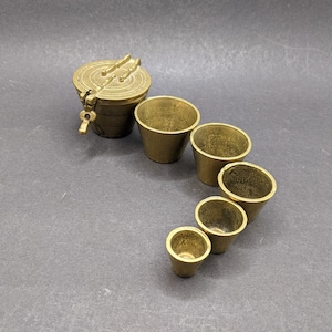 May include: A set of antique brass measuring cups and a small container with a hinged lid. The cups are graduated in size, and the container has a handle and a latch. The items are arranged on a dark grey surface.