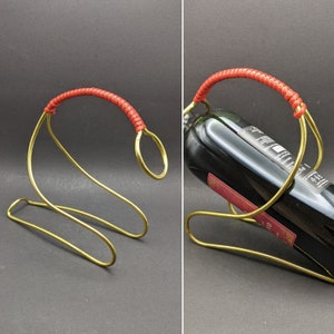 May include: A gold-colored metal wine bottle holder with a red handle. The holder has a curved design and is holding a dark wine bottle. The handle is wrapped in a textured red material.