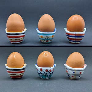 May include: Six ceramic egg cups with colorful patterns. Each cup holds a brown egg. The cups have a variety of designs, including stripes, polka dots, and floral patterns.
