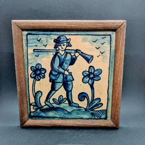 Könnte beinhalten: Eine gerahmte Fliese mit einem blauen und weißen bemalten Design einer Person, die ein Gewehr trägt und durch ein Feld mit Blumen geht. Der Hintergrund ist hellgelb.