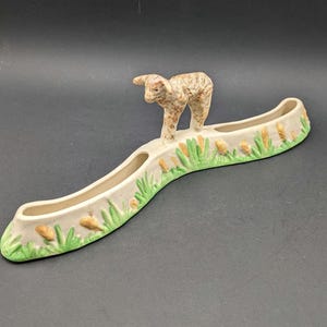 May include: A ceramic lamb figurine stands atop a cream-colored, curved planter with two openings. The planter is decorated with green grass and tan cattails. The lamb is tan and brown.