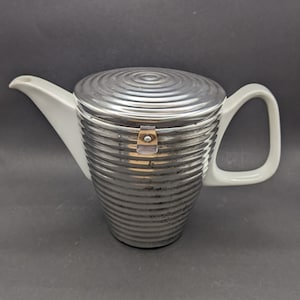 May include: A white ceramic teapot with a silver lid and a silver handle. The lid has a circular pattern and a silver latch.