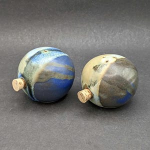 May include: Two spherical ceramic bottle stoppers. One is blue with lighter blue and tan streaks. The other is tan, brown, and blue. Both have cork stoppers.