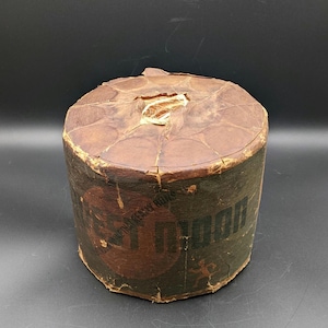 May include: Cylindrical container with a brown, aged appearance. The top is made of circular pieces of brown material. The side of the container has the words "ST. DIDON" in black and red lettering. The container appears to be made of cardboard.