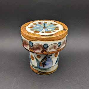 Puede incluir: Una caja de baratijas de cerámica con una tapa que presenta un diseño radial en azul, verde y marrón. La caja tiene una base cilíndrica con un pájaro azul pintado y detalles florales. El borde es de color marrón cálido.