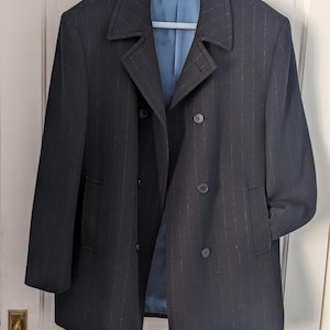 以下が含まれることがあります： 薄いストライプが入った濃いネイビーのダブルブレストコート。水色のハンガーにかかっています。裏地は水色で、クラシックなノッチドラペルカラーです。フロントポケットが2つ、ボタンが3つ付いています。