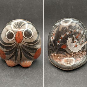 May include: Two rounded ceramic objects featuring hand-painted designs. The left object is a stylized owl with black, gray, and orange details. The right object is a smooth, flat-faced stone with a painted scene of a bird among foliage.