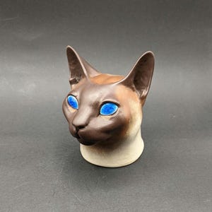 Puede incluir: Una figura de cerámica con forma de cabeza de gato que presenta una cara y orejas de color marrón oscuro que se transforman en un cuello de color crema más claro. El gato tiene unos llamativos ojos azules brillantes en forma de almendra.